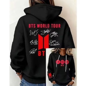 Può includere: Una felpa nera con cappuccio caratterizzata da un logo BTS rosso e firme bianche sul retro. La parte anteriore mostra una grafica rossa con tre cerchi e la scritta BTS, insieme a cordini bianchi e un logo rosso sul polsino della manica.