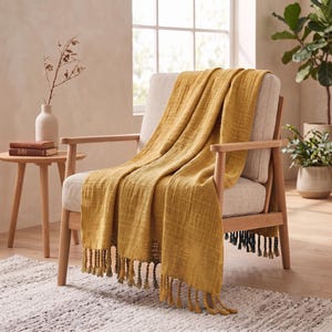 Può includere: Una coperta color giallo senape con una finitura in tessuto testurizzato e stropicciato, caratterizzata da frange annodate lungo il bordo inferiore. La coperta è drappeggiata su una poltrona in legno chiaro con seduta e schienale imbottiti.
