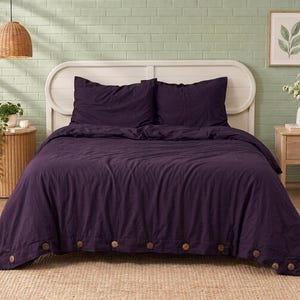 Könnte beinhalten: Ein tiefviolettes Bettbezug-Set mit zwei passenden Kissenbezügen und einer Reihe großer, runder, hellbrauner Holzknöpfe an der Unterkante. Die Bettwäsche hat eine strukturierte, matte Oberfläche und liegt auf einem Bett mit weißem Kopfteil.