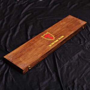 May include: A rectangular wooden storage box with a dark brown wood grain finish. The lid features a gold-outlined shield emblem with a red cross inside and the gold text "Non Nobis Domine Non Nobis" printed below it. The box has gold metal latches.