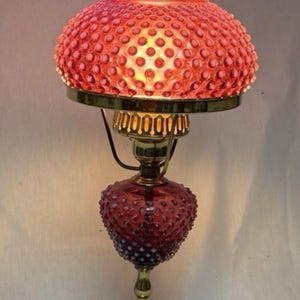 Könnte beinhalten: Eine Tischlampe mit einem rosafarbenen Hobnail-Glasschirm mit gewelltem Rand und einem passenden rosafarbenen Hobnail-Glasfuß. Die Lampe verfügt über einen messingfarbenen Metallständer und einen quadratischen weißen Marmorsockel mit Licht.