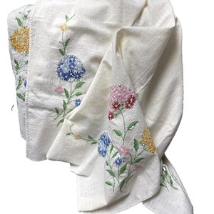 May include: An off-white textile featuring embroidered floral motifs in shades of yellow, blue, pink, and red. The fabric has a visible woven texture, and the embroidery depicts various flower stems with green leaves arranged across the surface.