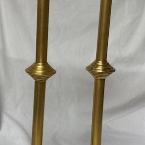 May include: A pair of tall, polished gold-colored metal candlesticks. Each candlestick features a wide, circular base, a slender vertical stem with a central decorative ribbed ring, and a flared, circular top platform for holding a candle.