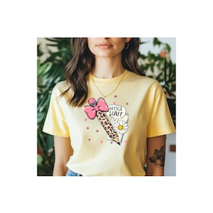 May include: A light yellow t-shirt featuring a graphic print of a pencil with a leopard print pattern, a pink bow, a white daisy, and a notepad with the text OFFICE STAFF, accented by small pink star-shaped icons scattered around the design.
