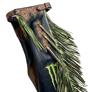 May include: Black leather chaps featuring a tan floral-tooled leather waistband and side panels. The chaps are adorned with long, metallic lime green fringe, three vertical brass-toned metal accents, and a bright lime green claw-mark graphic.