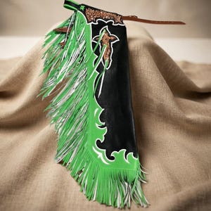 Pode incluir: Uma peça de perna de chaparreras em couro preto com sobreposições de couro verde brilhante e franjas longas. O design inclui um intrincado trabalho floral na seção superior e um detalhe recortado em forma de cruz. Uma tira de couro marrom.