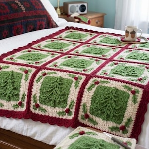 May include: A handmade crochet blanket featuring a grid of square panels, each displaying a textured green Christmas tree on a cream background, bordered by dark red yarn with small red berry accents and a scalloped edge finish.