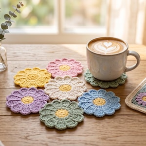 May include: A collection of six round, flower-shaped crochet coasters in pastel yellow, pink, white, purple, sage green, and light blue, alongside a square crochet potholder featuring a floral pattern and a ceramic mug filled with latte art.