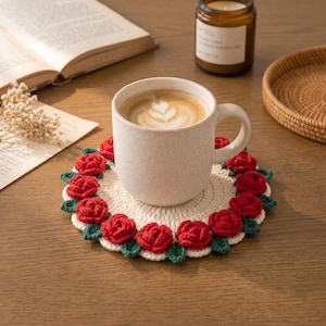 Könnte beinhalten: Ein runder, cremefarbener Häkeluntersetzer mit einem dekorativen Rand aus kleinen, leuchtend roten Wollrosen und dunkelgrünen Blättern. Eine gesprenkelte, helle Keramiktasse mit Kaffee steht mittig auf dem strukturierten Untersetzer.