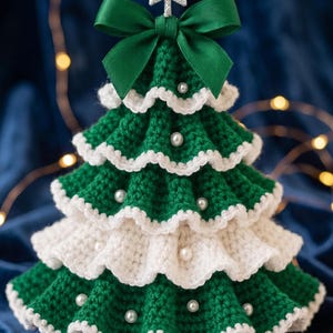 Puede incluir: Una decoración de árbol de Navidad tejida a ganchillo con volantes escalonados en verde y blanco. El árbol está adornado con pequeñas perlas blancas y rematado con un lazo de cinta de satén verde oscuro y una estrella plateada brillante.