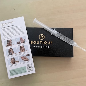 Op de afbeelding: Een Boutique Whitening set met een zwarte rechthoekige doos met gouden logo, een doorzichtige plastic spuit gevuld met 16% carbamideperoxide tandenbleekgel en een witte instructiefolder met stapsgewijze diagrammen en tekst.