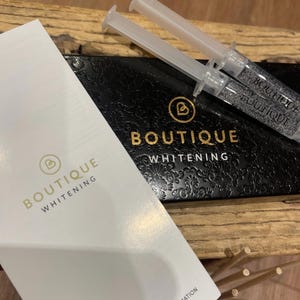 May include: Two clear plastic syringes filled with gel rest on a black box with gold text reading BOUTIQUE WHITENING. A white rectangular card with the same gold logo and text sits in the foreground, featuring the words INSTRUCTIONS AND COSHH INFORMATION.