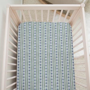 May include: A light pink wooden crib featuring a fitted sheet with a repeating vertical pattern. The sheet design consists of alternating stripes of green, white, and blue, with a small blue floral motif centered within the blue vertical bands.
