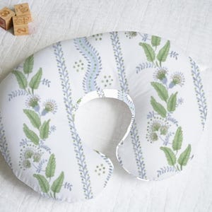 May include: A white and blue patterned nursing pillow with a green floral design. The pillow is shaped like a horseshoe and has a hole in the center for the baby's head.