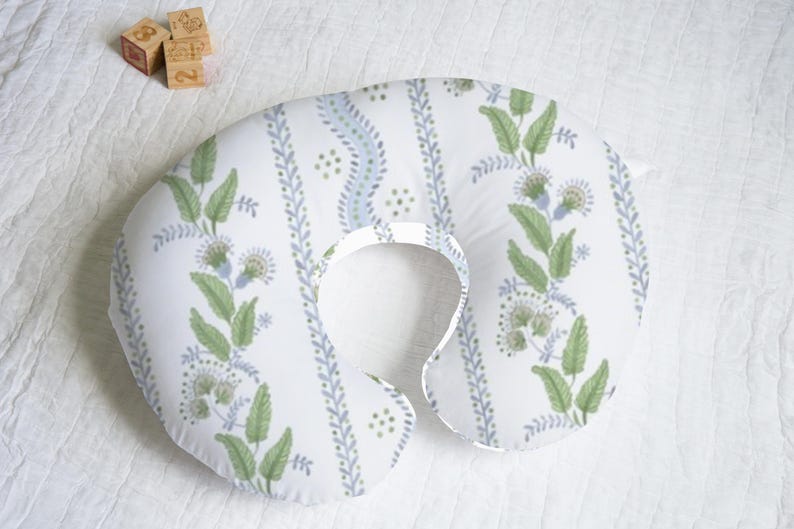 May include: A white and blue patterned nursing pillow with a green floral design. The pillow is shaped like a horseshoe and has a hole in the center for the baby's head.