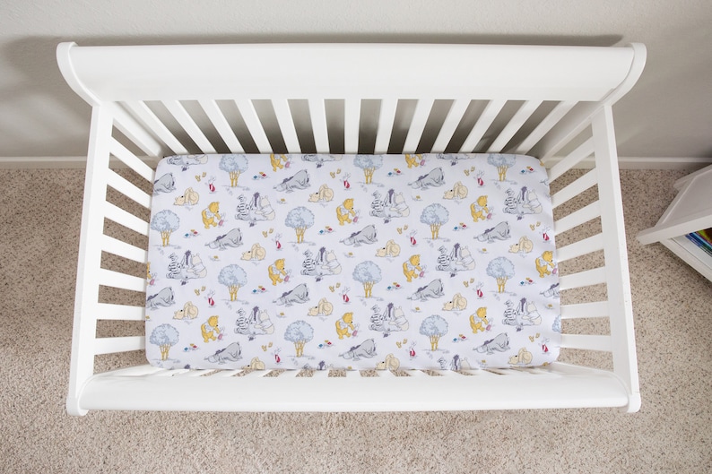 May include: A white cot with a fitted sheet featuring a Winnie the Pooh and friends print. The sheet has a light blue background with yellow, grey, and brown characters.