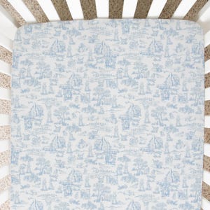 Puede incluir: Una sábana de cuna blanca con un patrón de toile azul y blanco. La sábana es ajustable y tiene una textura suave y lisa.