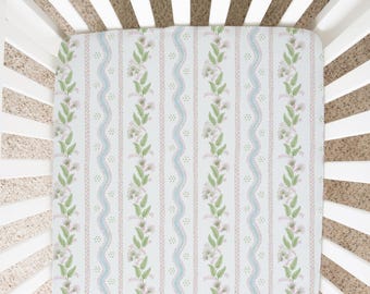 May include: A white cot with a fitted sheet featuring a floral pattern in shades of pink, green, and blue. The pattern includes small flowers and leaves, and a wavy line design.