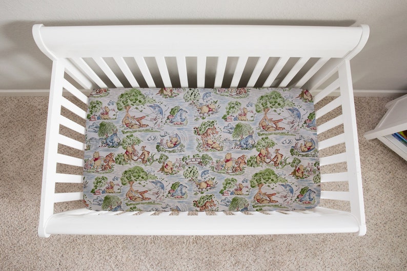 May include: White cot with a Winnie the Pooh themed fitted sheet. The sheet features a light blue background with a repeating pattern of Winnie the Pooh and his friends in a woodland setting.