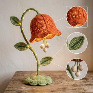 Puede incluir: Una lámpara de flor de ganchillo con un tallo verde curvado, una gran pantalla naranja en forma de campana y tres pequeñas esferas de color crema colgando del centro. El tallo tiene hojas verdes y luces integradas sobre una base en forma de flor.