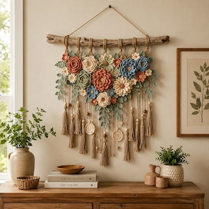 Op de afbeelding: Een wandhanger met gehaakte bloemen in tinten terracotta, saliegroen, crème en stoffig blauw, bevestigd aan een stuk drijfhout. Het arrangement bevat hangende garenkwastjes, houten kralen en cirkelvormige kanten accenten als decoratie.
