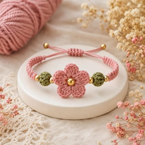 May include: A pink crocheted flower bracelet featuring a central gold-toned bead, two olive green crocheted spherical beads, and smaller gold-toned accent beads. The bracelet has a pink cord band with an adjustable sliding knot closure mechanism.