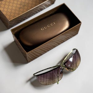 May include: Gucci sunglasses with thin gold-tone metal frames and gradient lenses, accompanied by a brown hard-shell case with the Gucci logo in gold lettering and a matching brown box featuring a repeating monogram pattern on the interior lid.