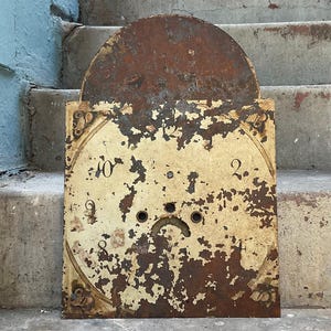 May include: An antique clock face with a square base and a semi-circular top. The face is off-white with peeling paint, revealing the metal underneath. Numbers 2, 8, 9, and 10 are visible. The top is heavily rusted.
