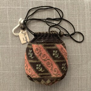 May include: A small, hand-stitched pouch with a drawstring closure. The pouch is primarily brown with diagonal stripes of pink and green. A tag with handwritten text is attached to the drawstring. The pouch is likely a vintage or antique item.