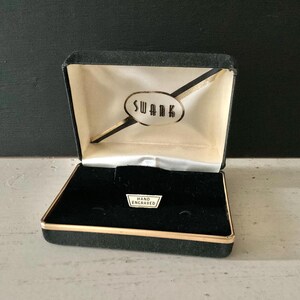 May include: Open black velvet jewelry box with gold trim. The interior features a black velvet base and a cream-colored lid with the word "SWANK" in gold lettering. A small tag reads "HAND ENGRAVED".