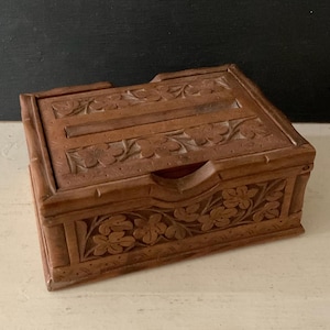 May include: A rectangular wooden box with intricate floral carvings. The box is a warm brown color and features detailed relief patterns of flowers and leaves on the lid and sides. The box has a small opening on the front.