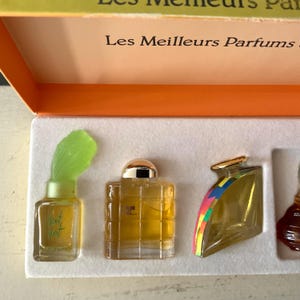 May include: A collection of miniature perfume bottles in a box. The bottles are different shapes and sizes, filled with a golden liquid. One bottle has a green, leaf-shaped top. The box is orange and white, with French text.