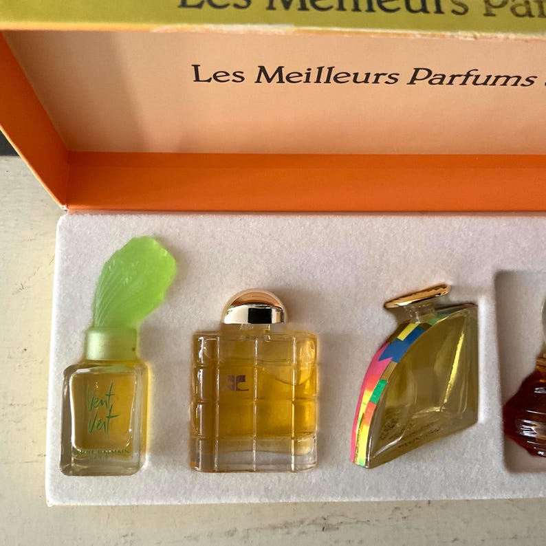 May include: A collection of miniature perfume bottles in a box. The bottles are different shapes and sizes, filled with a golden liquid. One bottle has a green, leaf-shaped top. The box is orange and white, with French text.