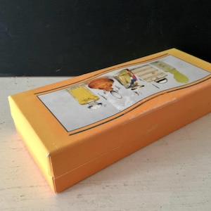 May include: A closed orange rectangular box with a decorative lid. The lid features an illustration of perfume bottles in yellow, gold, and brown. The box is new and appears to be unused.