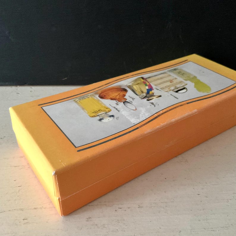 May include: A closed orange rectangular box with a decorative lid. The lid features an illustration of perfume bottles in yellow, gold, and brown. The box is new and appears to be unused.