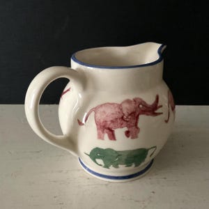 May include: Cream-colored ceramic pitcher with a rounded body and a curved handle. The pitcher is decorated with pink and green elephant illustrations. A blue stripe accents the base and rim.