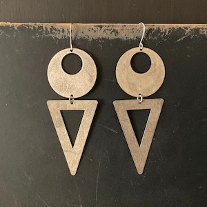 May include: A pair of geometric earrings with a circular top and a triangular bottom. The earrings are made of a brushed, metallic material with a silver hook. The design is modern and minimalist, with a vintage aesthetic.