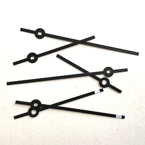 May include: Several black watch hands are scattered on a white surface. The hands have a simple, minimalist design with a circular or rectangular detail at one end and a straight, elongated shape. Some hands have a small white rectangle at the tip.