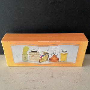 May include: An orange box with a white and grey border displaying five miniature perfume bottles. The bottles are different shapes and colours, including yellow, brown, and clear. The box is on a white surface.
