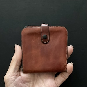 May include: A square-shaped brown leather wallet featuring a smooth texture and a central vertical strap closure secured with a single black snap button. The wallet has rounded corners and visible stitching along the edges of the strap and body.