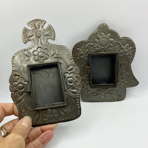 May include: Two decorative metal frames with a dull grey finish, featuring embossed floral patterns and scalloped edges. Each frame has a central rectangular opening and a hinged side mechanism, showcasing intricate metalwork and a weathered texture.