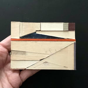 May include: A small, rectangular art piece composed of layered, geometric shapes in neutral tones, accented with a vibrant orange stripe. The artwork features a mix of textures and colors, including navy blue and a touch of burgundy.