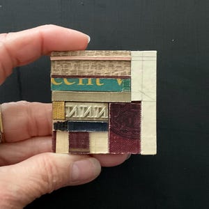 May include: A small, square collage composed of various textured materials. The collage features strips of fabric, paper, and other elements in shades of beige, green, gold, and burgundy. The word "IIII" is visible in a metallic font.