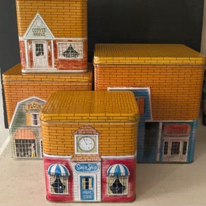 May include: A collection of vintage tin boxes, each designed to resemble a different shop. The boxes feature brickwork patterns and are painted with details like a coffee house, flour shop, and sugar shop. The lids are yellow.