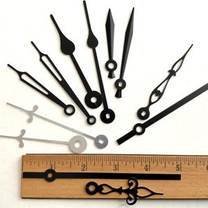 May include: A collection of black and silver clock hands in various styles, including pointed, teardrop, and arrow shapes. A wooden ruler with inch markings is also present, providing scale. The hands are designed for clock making and repair.