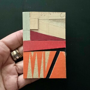 May include: Abstract mixed media art piece with geometric shapes. The artwork features a rectangular composition with layers of red, orange, beige, and black elements. The piece is held in a hand, showcasing the texture and color variations.