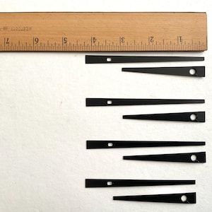 May include: A collection of black clock hands in various sizes, with a wooden ruler in the background. The hands have a rectangular or triangular shape with a small hole or square near the end. The ruler is marked in inches.