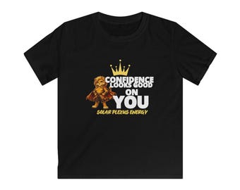 Puede incluir: Una camiseta negra de manga corta con un gráfico de un león con capa y corona. El texto dice "CONFIDENCE LOOKS GOOD ON YOU" en blanco y "SOLAR PLEXUS ENERGY" en amarillo, acompañado de un pequeño icono de corona amarilla arriba.