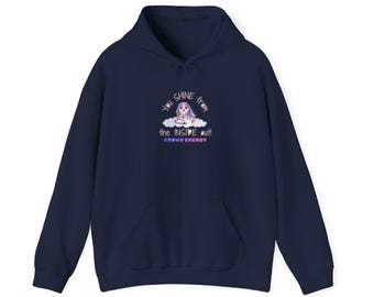 Puede incluir: Una sudadera con capucha azul marino que presenta un gráfico de una pequeña criatura de tonos morados sentada sobre una nube. El texto dice: "YOU SHINE from the INSIDE out!" con las palabras "CROWN ENERGY" impresas en azul y rosa.