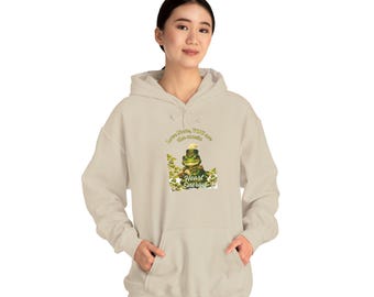 Puede incluir: Una sudadera con capucha de color beige que presenta un gráfico de una rana verde con sombrero tocando la flauta. El texto superior dice Love Note, YOU are the music, y el inferior Heart Energy, rodeado de hojas de color verde.
